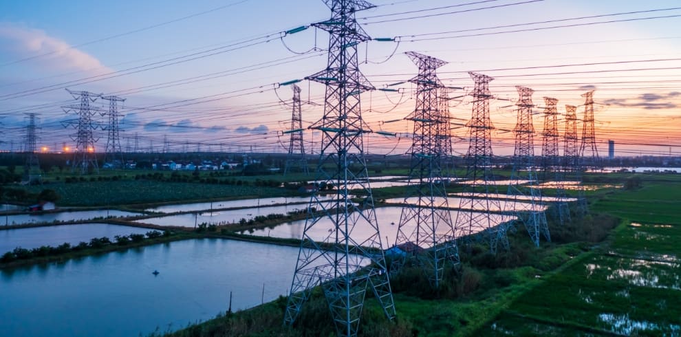 High-voltage power transmission lines and towers spanning a rural landscape with water channels at sunset, representing electric grid infrastructure, energy distribution, and sustainable power delivery in agricultural regions.