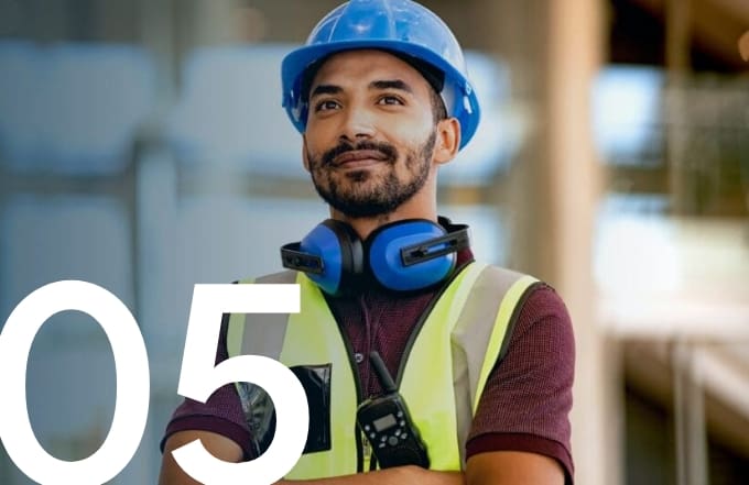 Construction worker wearing a safety vest, hard hat, and hearing protection, standing with arms crossed on a job site.