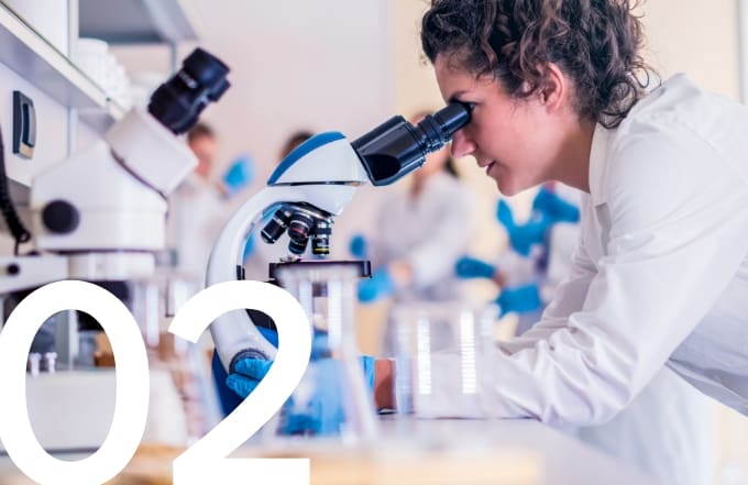 Female scientist in a laboratory using a microscope for research, surrounded by lab equipment and colleagues.