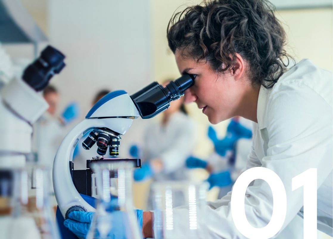 Female scientist in a laboratory using a microscope for research, surrounded by lab equipment and colleagues.