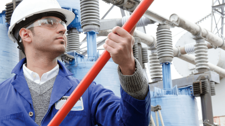 Engineer wearing a hard hat and safety glasses performing high-voltage equipment testing outdoors with an insulated hot stick.